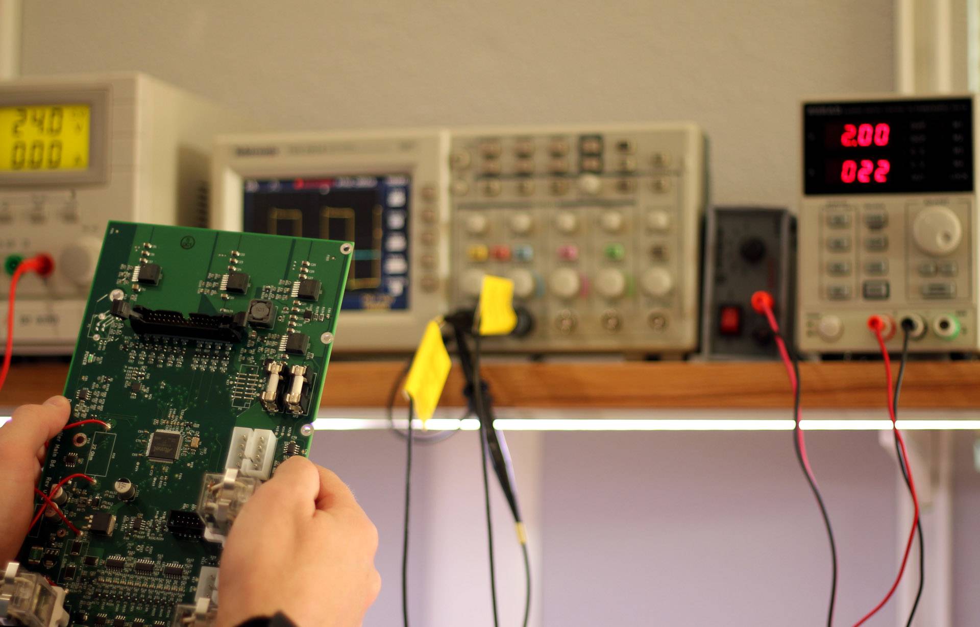 Hands holding a circuit board in front of a bench of test equipment